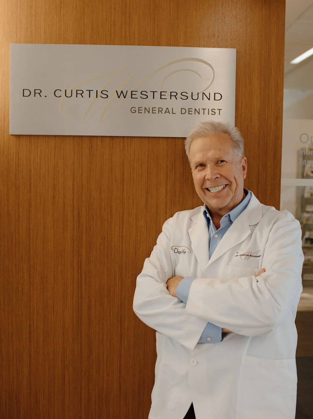 A smiling man in a white coat standing in front of a sign that says 'Dr. Curtis Westersund, General Dentist' in an office setting.