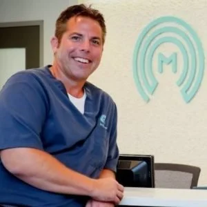Smiling man in blue scrubs sitting at a desk in an office with a logo on the wall