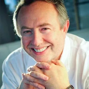 A smiling man with light hair and a white shirt, sitting with hands clasped, in a well-lit indoor setting.