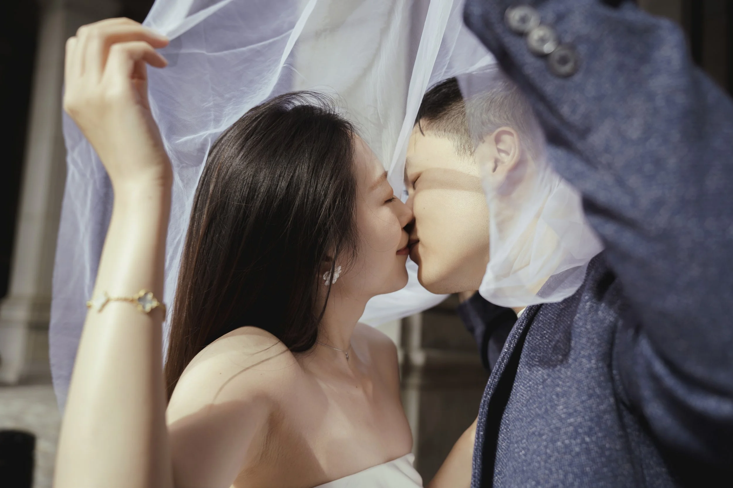 A couple sharing an intimate kiss behind a sheer white curtain, with the woman wearing a white dress and jewelry, and the man in a dark suit.