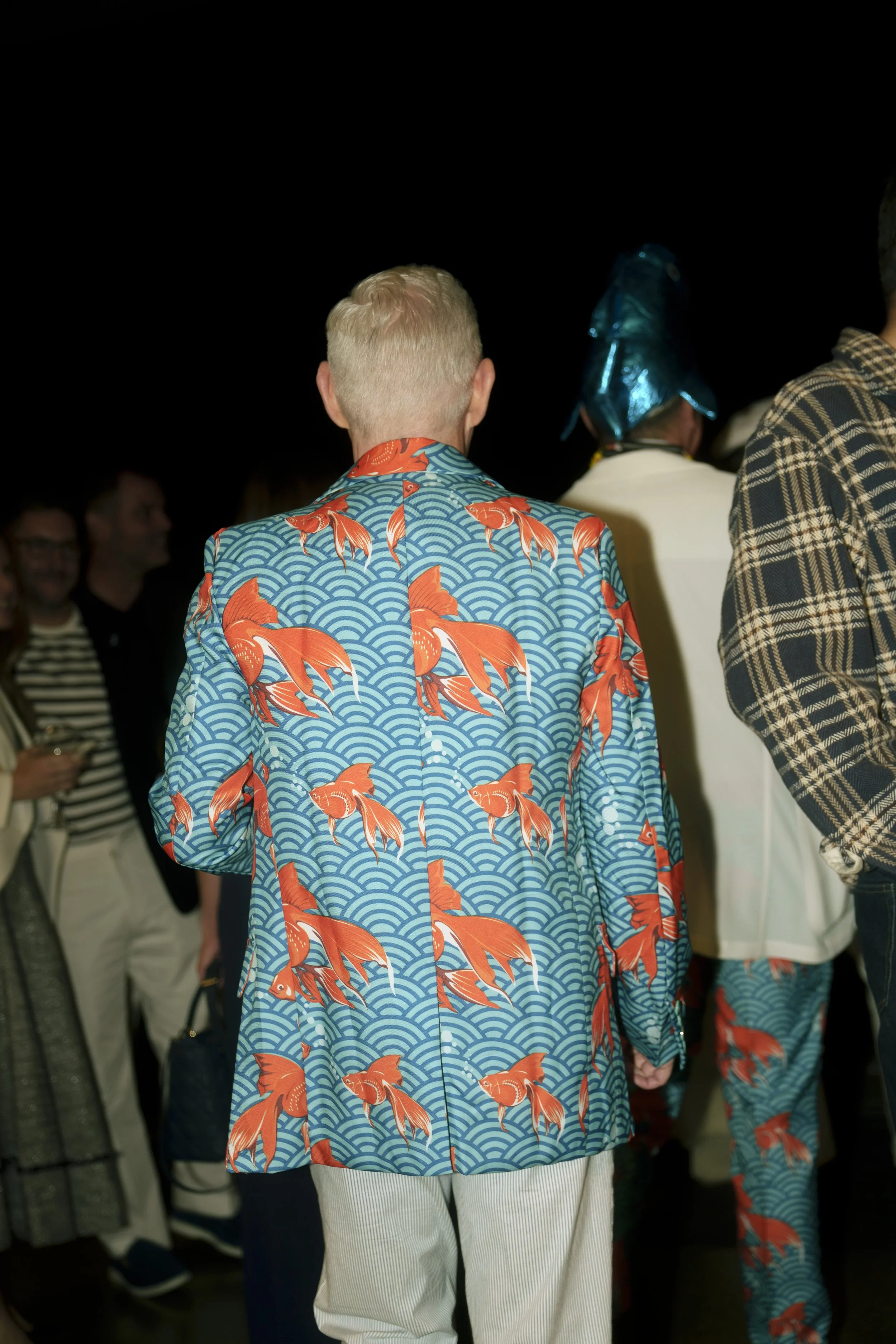 Back of a person in a colorful blazer with an orange fish pattern, attending an event among other people, some in eccentric attire like a metallic hat.