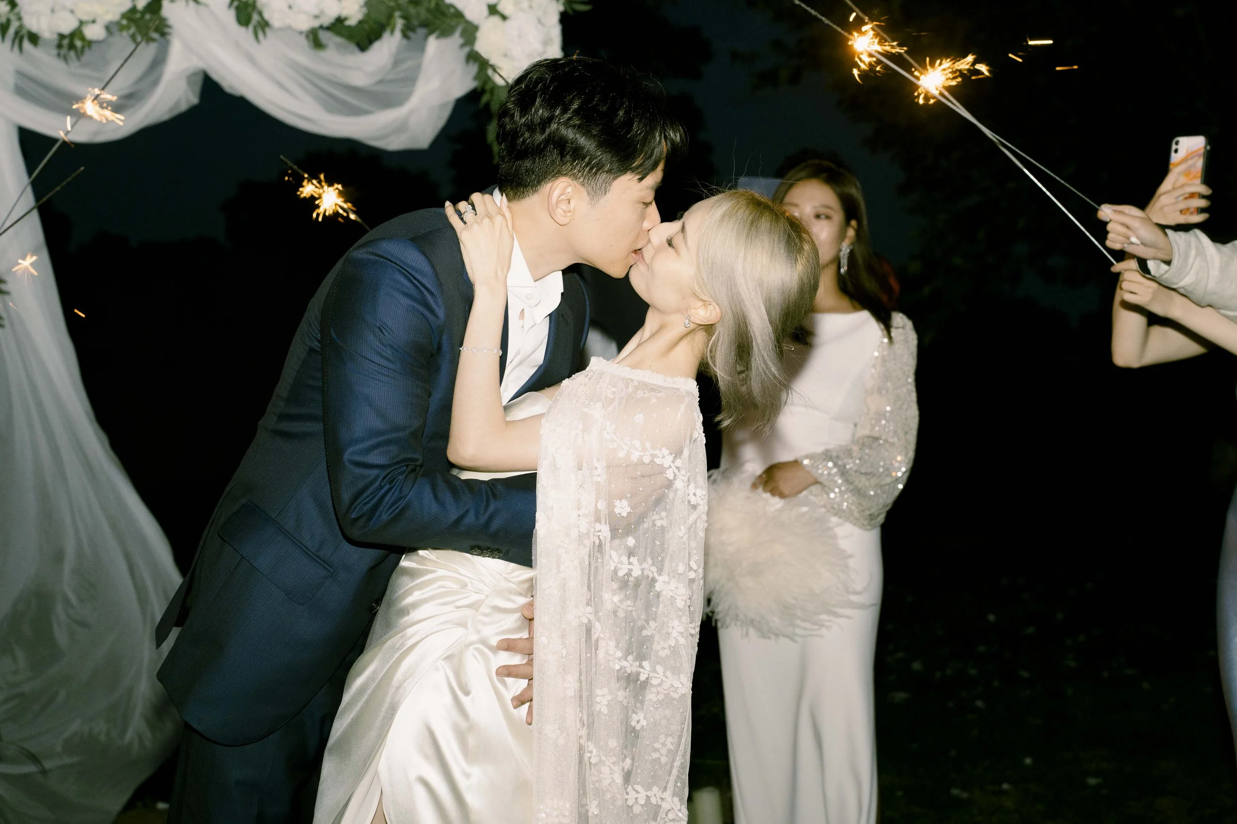 A couple, dressed in wedding attire, sharing a kiss during a wedding celebration at night, with guests holding sparklers in the background.