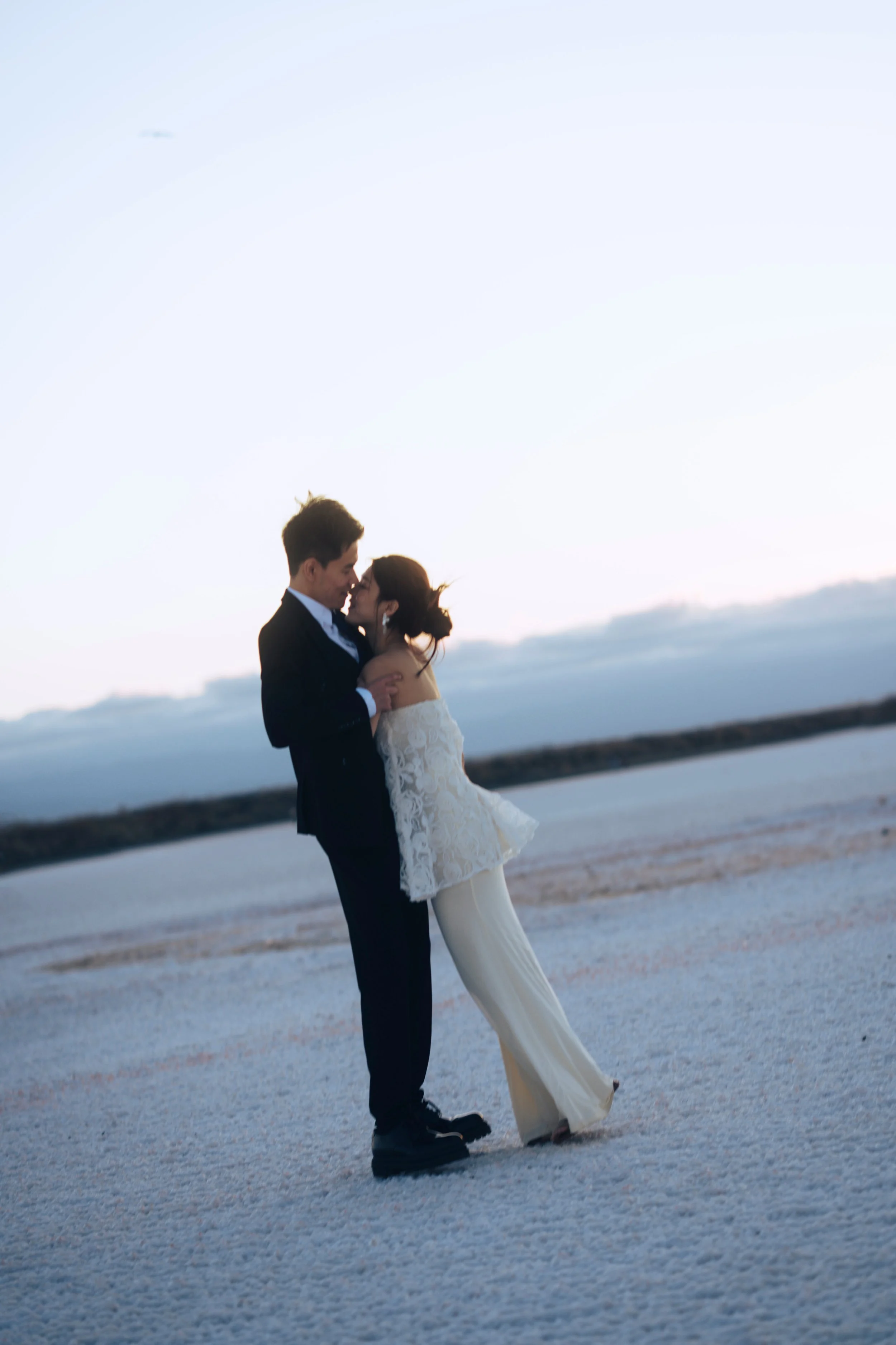 A couple dressed in wedding attire embracing on a beach at sunset.