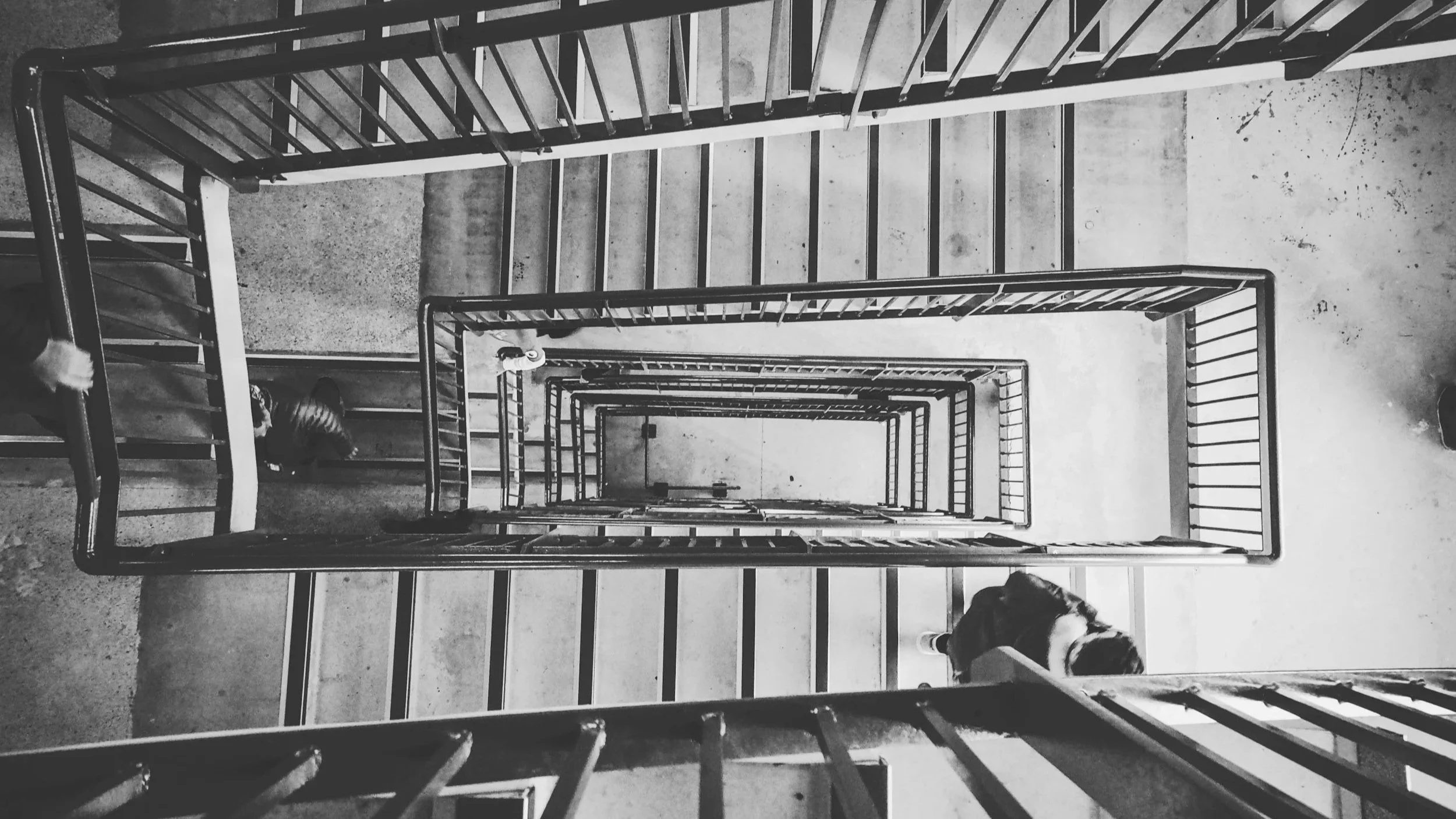 A black-and-white photograph taken from the top of a spiral staircase, looking down through the staircase's open center, showing multiple levels of the staircase and a concrete floor below.