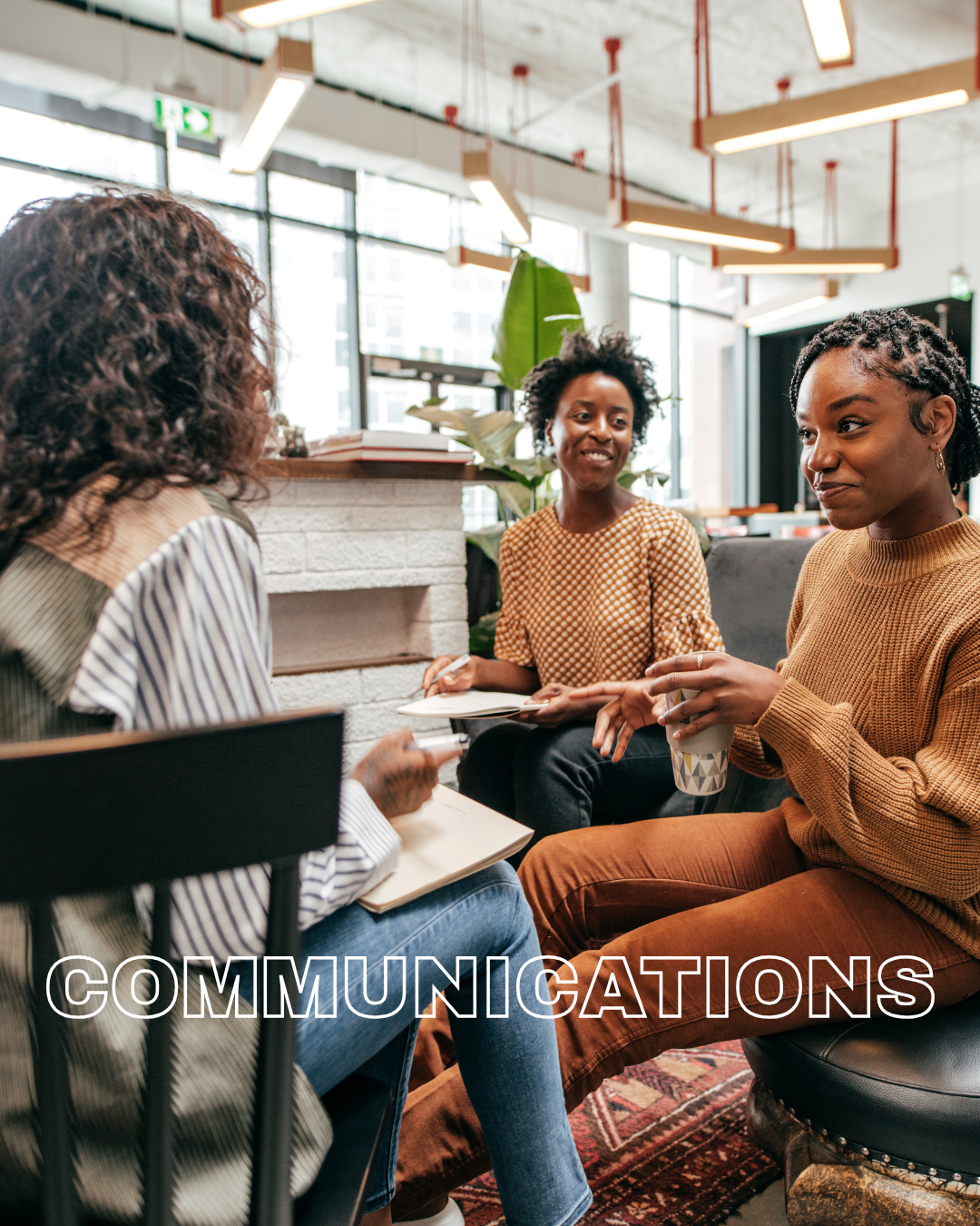 Three women having a conversation in a modern office or coffee shop, sitting on chairs around a small table. They are engaged and smiling, with one holding a notebook, another holding a coffee cup. The room has large windows, hanging lights, and indoor plants.