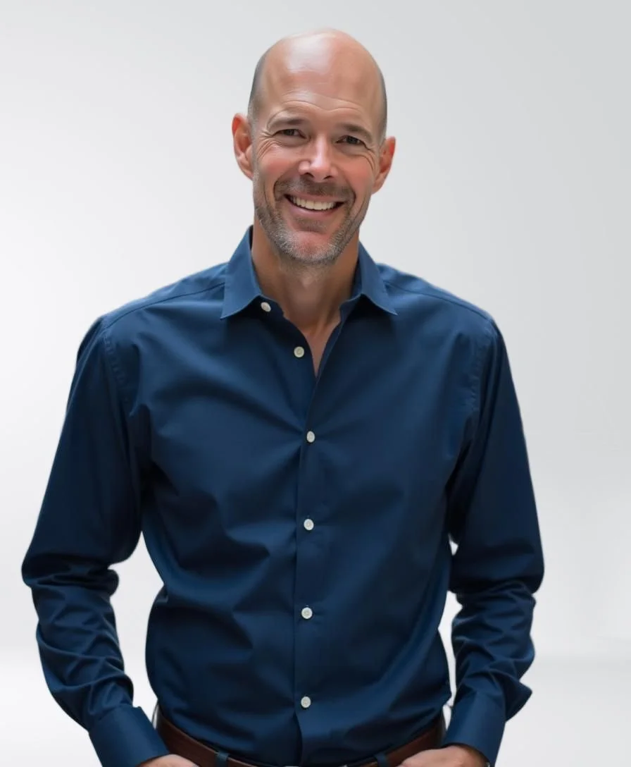 A middle-aged man with a bald head and gray beard, smiling, wearing a blue dress shirt, standing against a plain gray background.