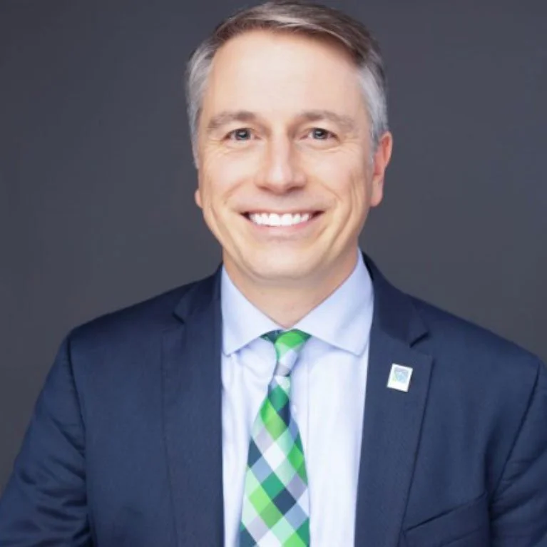 Smiling man in a dark blue suit, light blue shirt, green and white plaid tie, and with a small lapel pin, against a gray background.