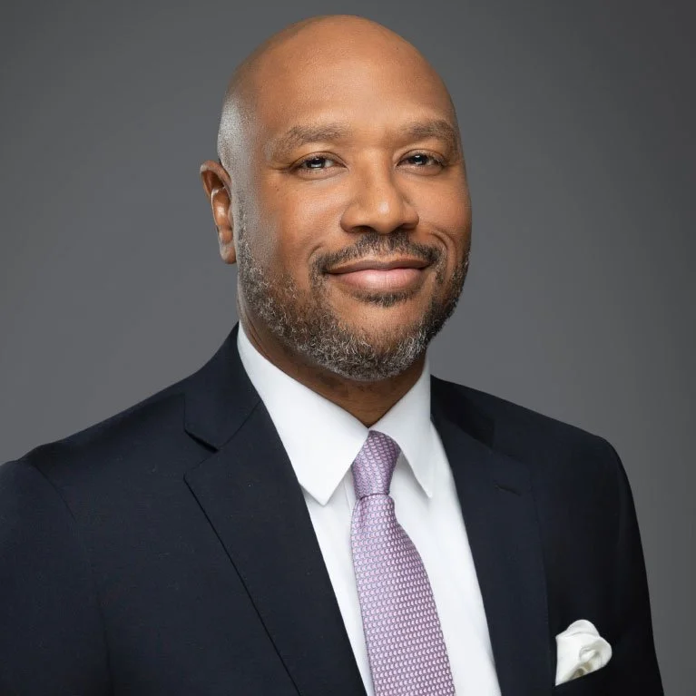 Professional headshot of a middle-aged African American man in a dark suit, white shirt, and light pink tie, smiling against a gray background.