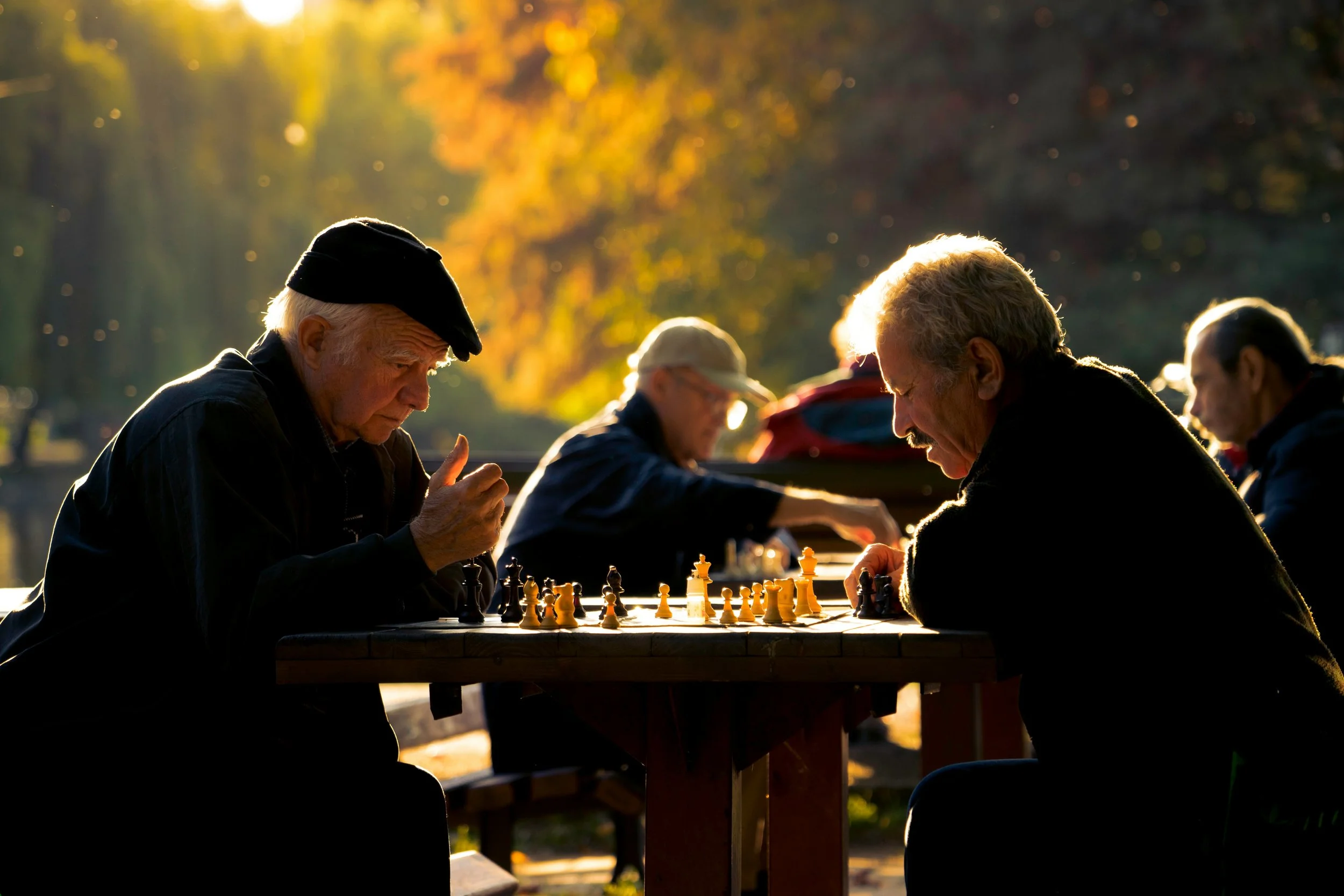 Older men playing chess outdoors during sunset with autumn trees in the background.