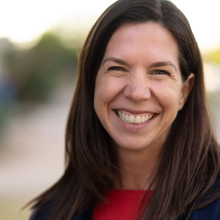 Close-up of a smiling woman with long dark hair and wearing a red top, outdoors with a blurred background.