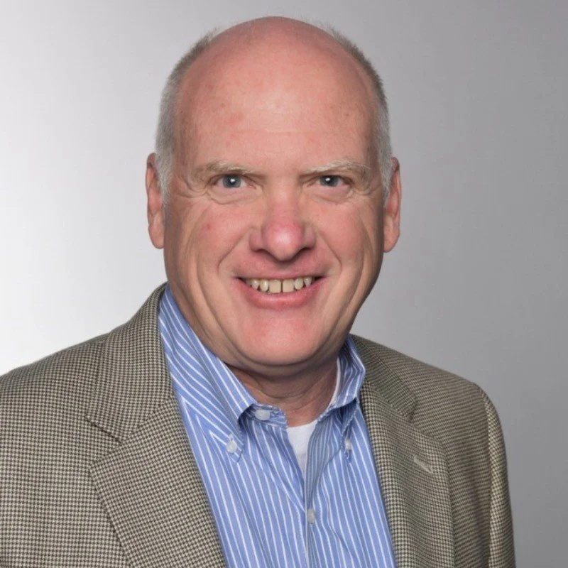 Headshot of a smiling middle-aged man wearing a checkered blazer and a blue striped dress shirt against a plain background.