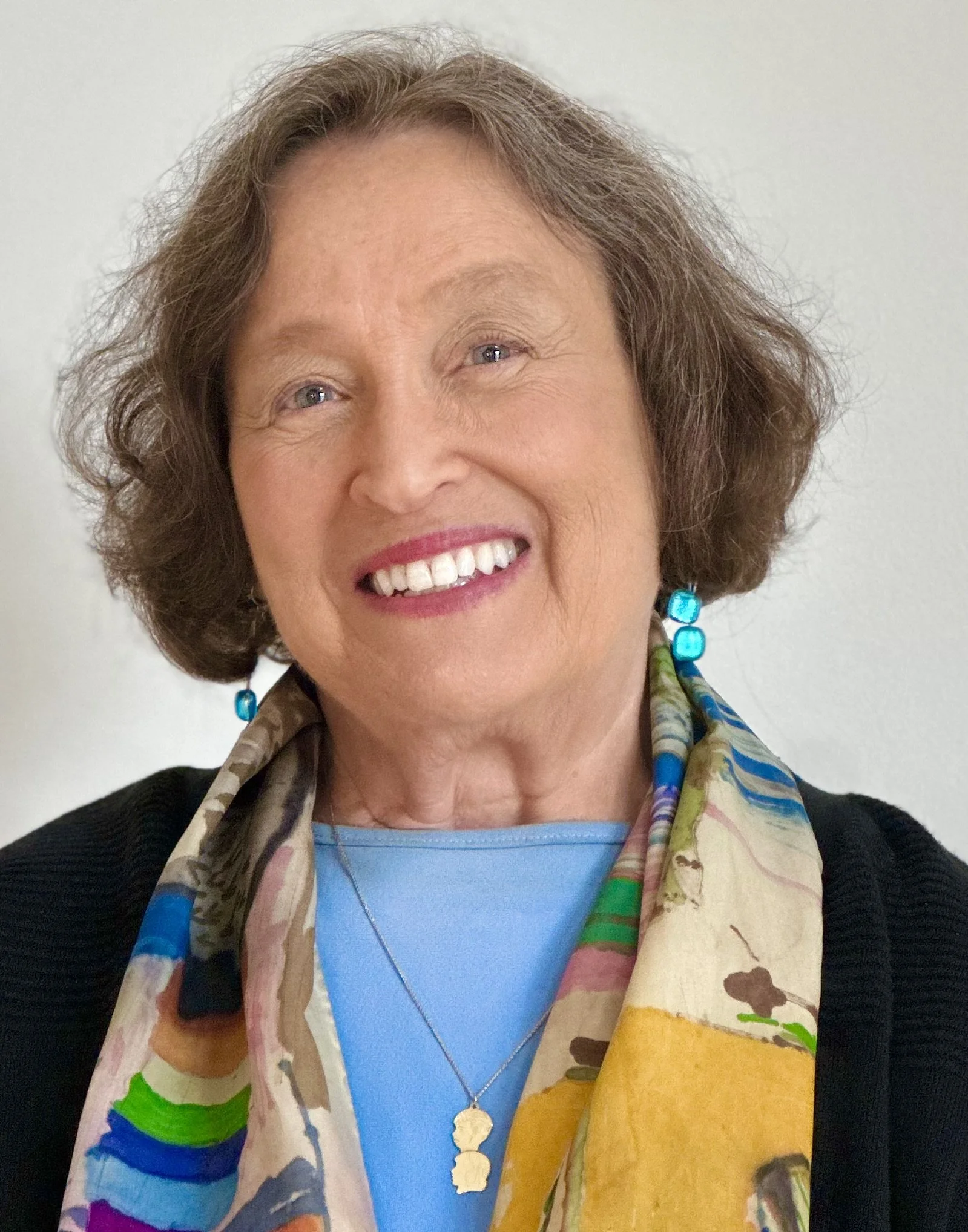A smiling elderly woman with short brown hair, wearing blue earrings, a colorful scarf, a blue top, and a black cardigan, facing the camera.
