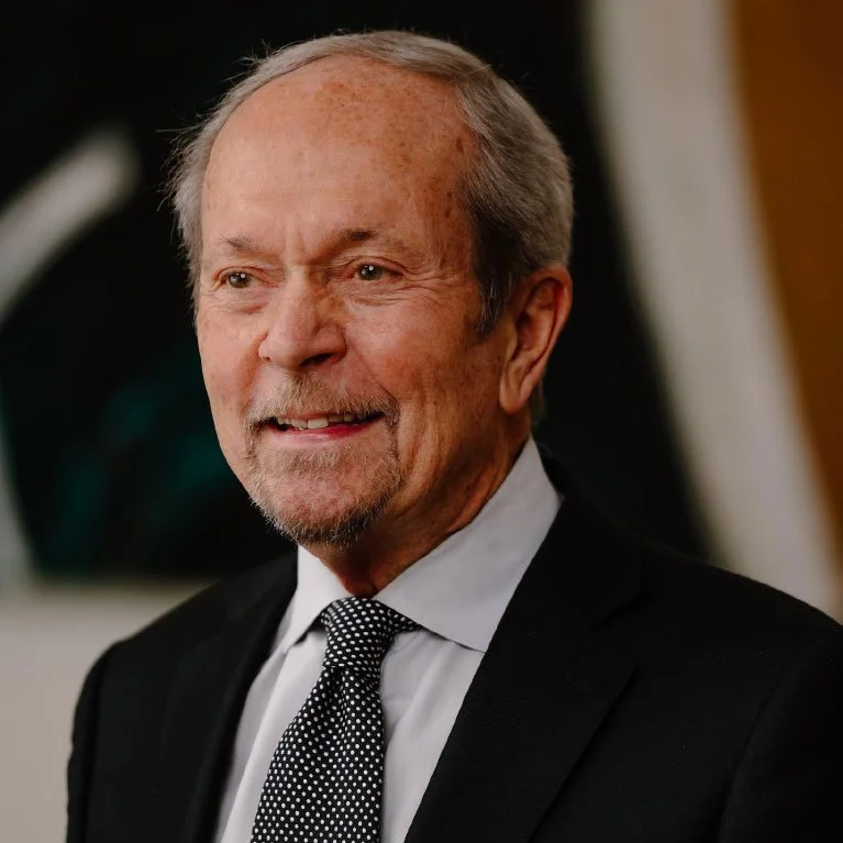 An older man with light skin, gray hair, and a beard, dressed in a black suit, white shirt, and black and white polka dot tie.