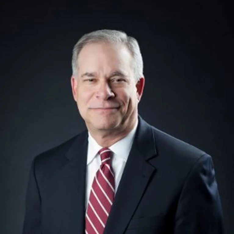 Professional headshot of a middle-aged man with gray hair, wearing a dark suit, white shirt, and red striped tie, standing against a dark background.