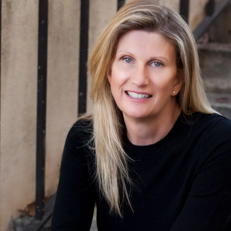 Portrait of a smiling woman with blonde hair wearing a black top, outdoors near a building with stairs.