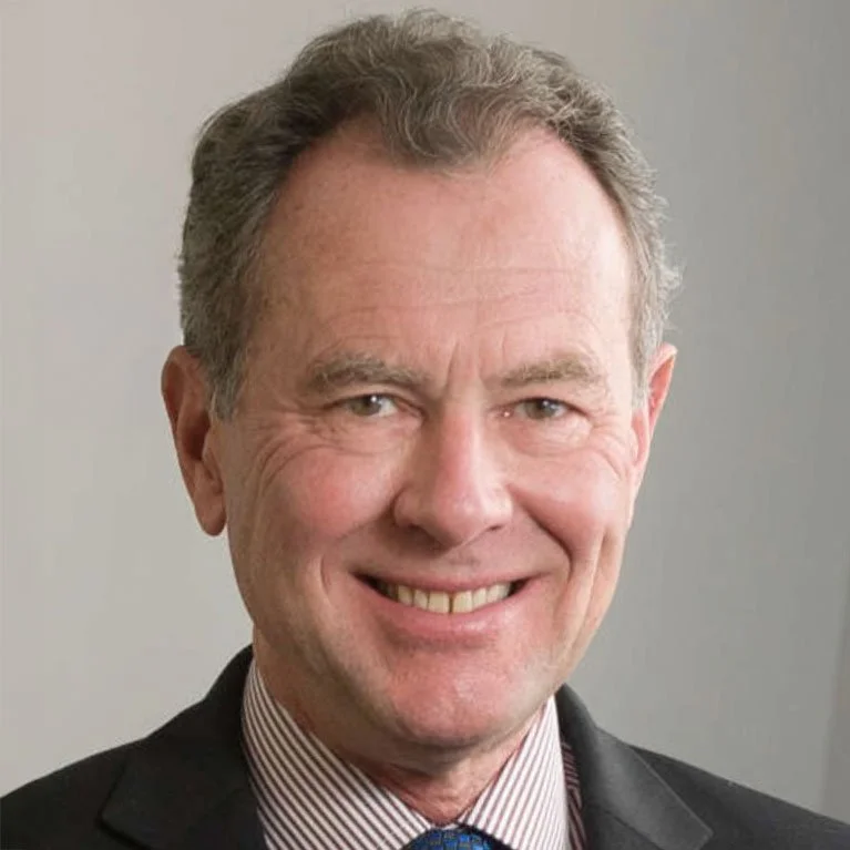 Close-up of a middle-aged man with light skin, gray hair, smiling, wearing a suit and tie.