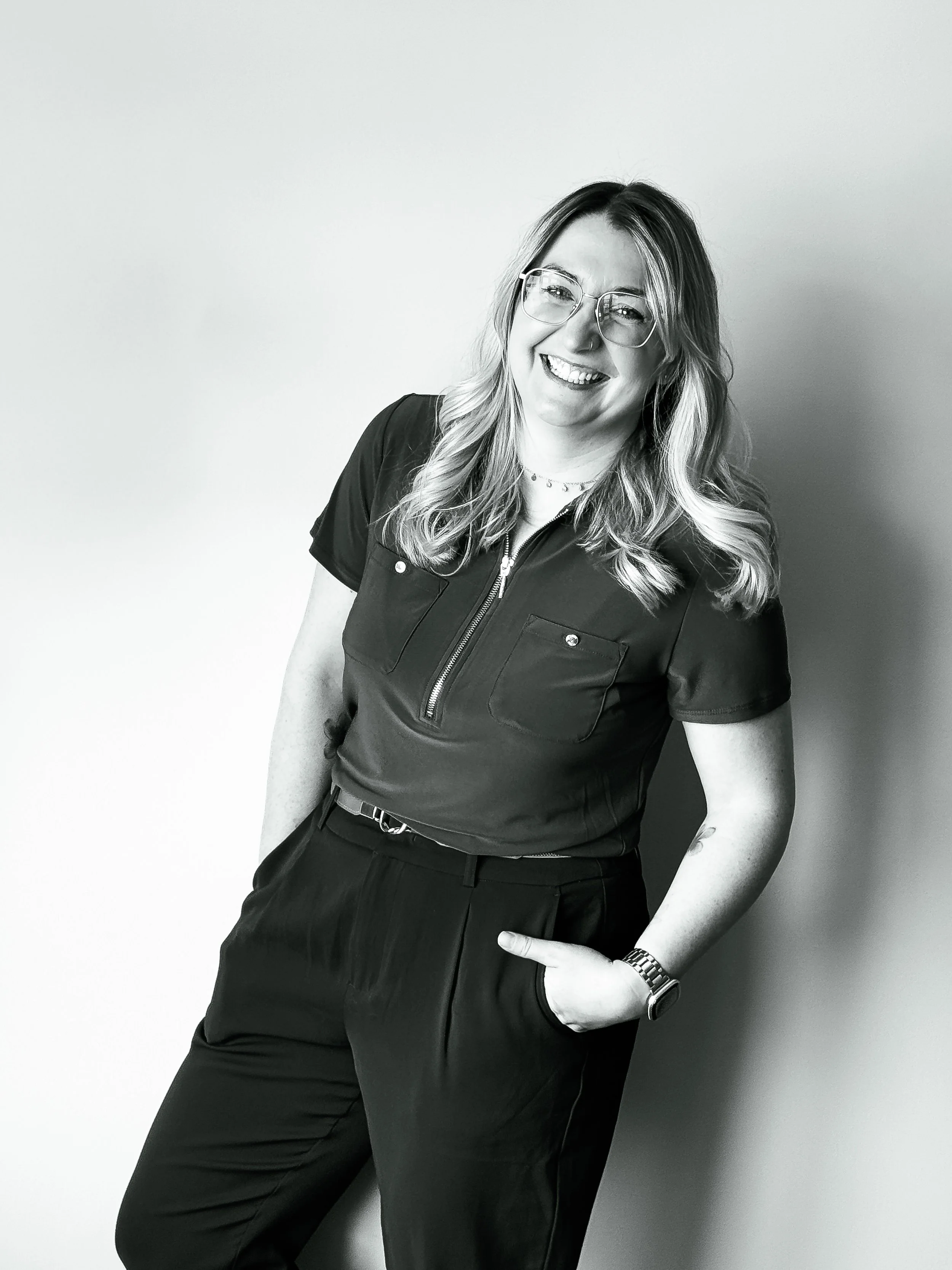 Black and white photo of a young woman with wavy hair, glasses, and a smile, standing against a plain wall, wearing a short-sleeved zip-up shirt and dark pants.
