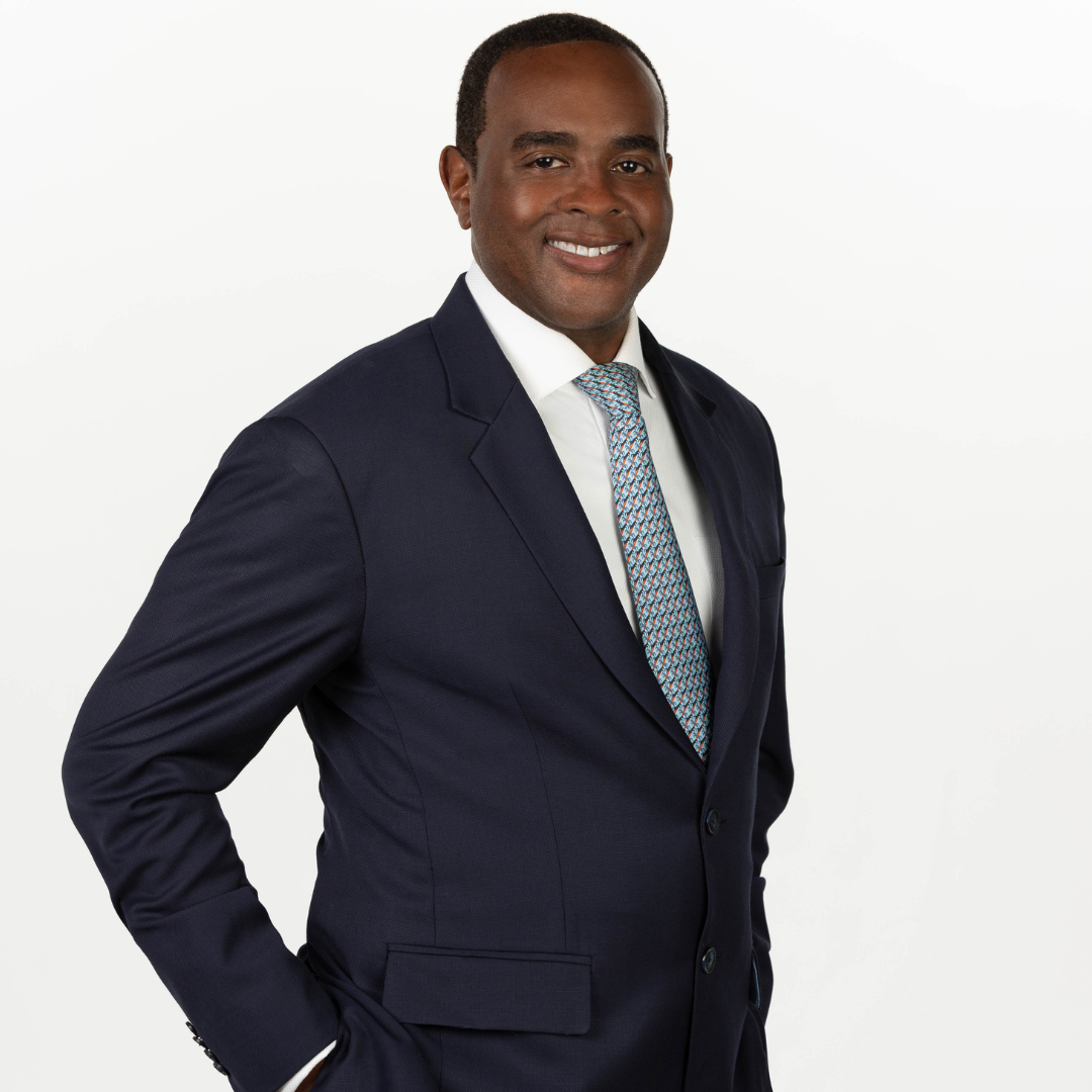 A man in a navy blue suit with a white shirt and patterned tie, smiling against a plain white background.