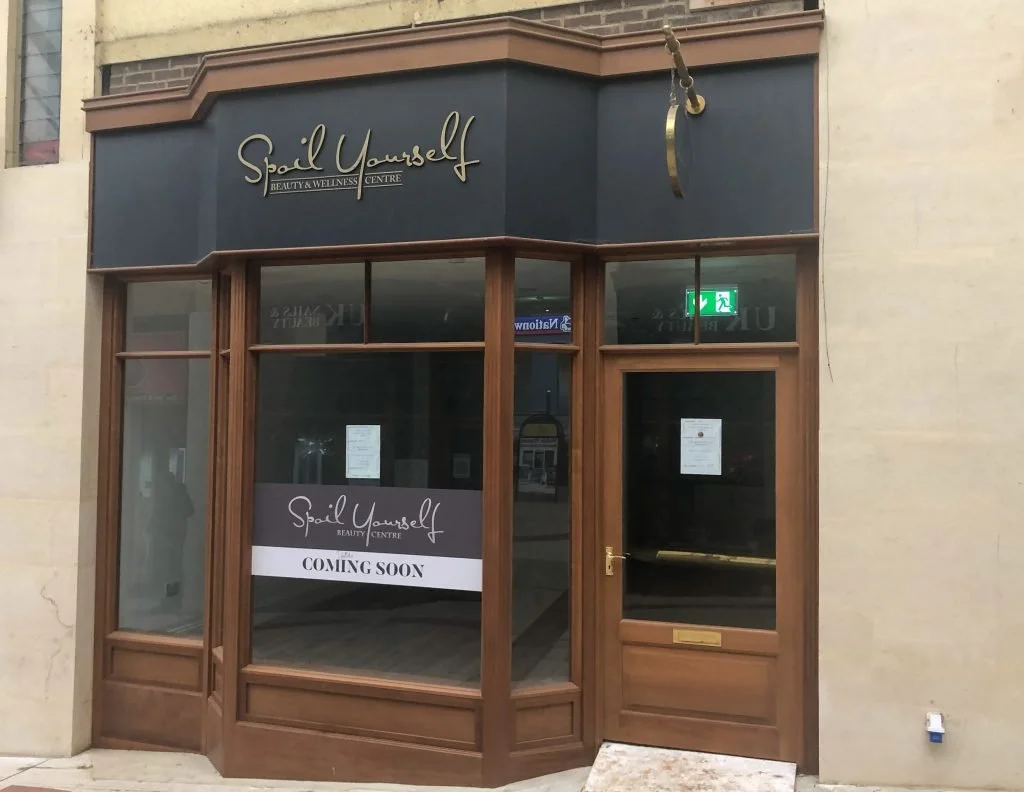 Front of a beauty and wellness center called 'Spail Yourself' with a sign indicating it is 'Coming Soon'. The storefront has large glass windows and wooden framing, with an emergency exit sign visible inside.
