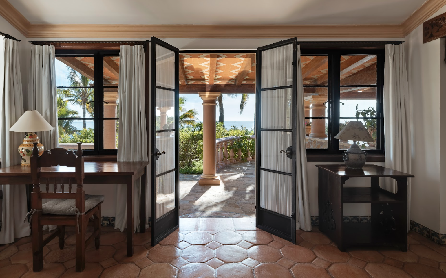Open French doors with black frames and white curtains reveal a sun-drenched terracotta patio with palm trees and ocean views at Casa Verde Norte, La Valise Los Cabos