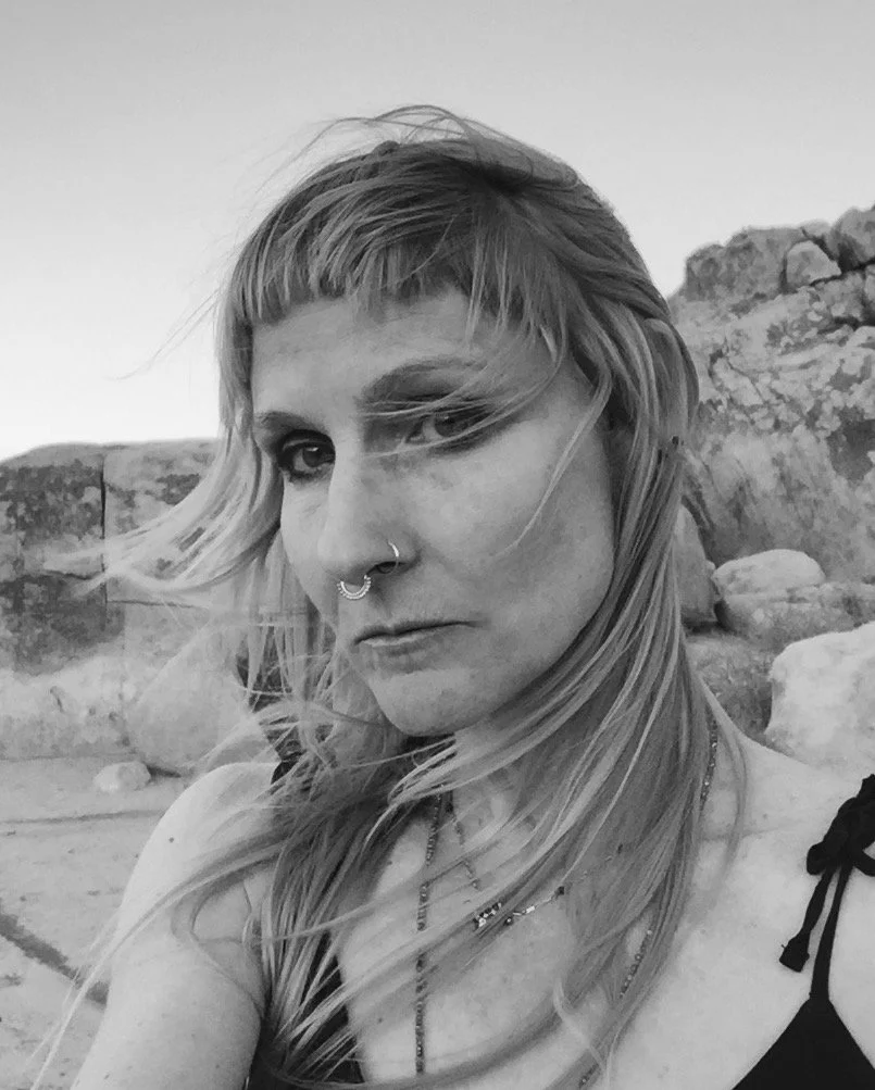 A woman with long blonde hair styled in space buns is touching her face and looking down, set against a dark background with warm sunlight.