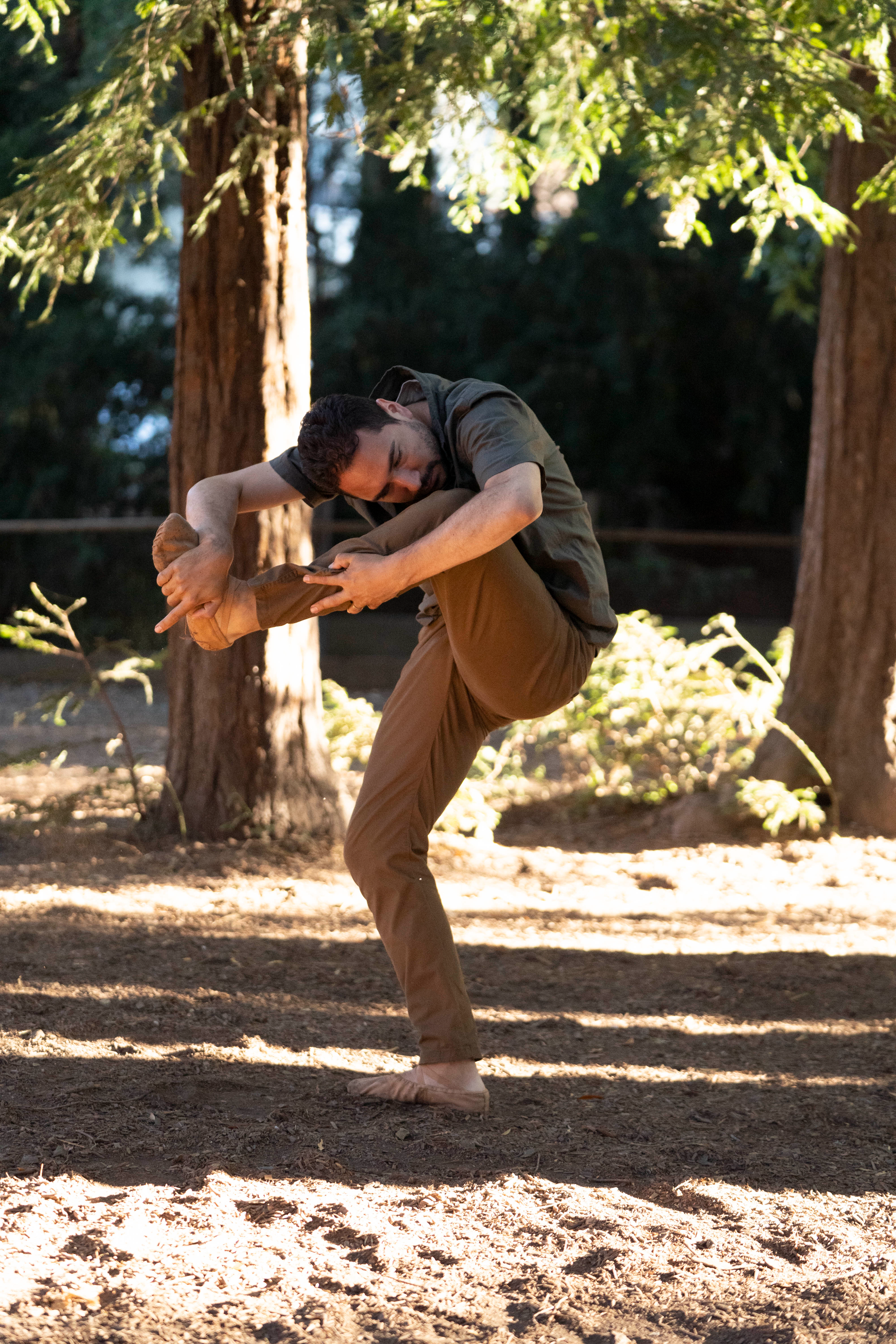 A man dancing outdoors in a wooded area, mid-move with one leg raised and arms gripping his foot.