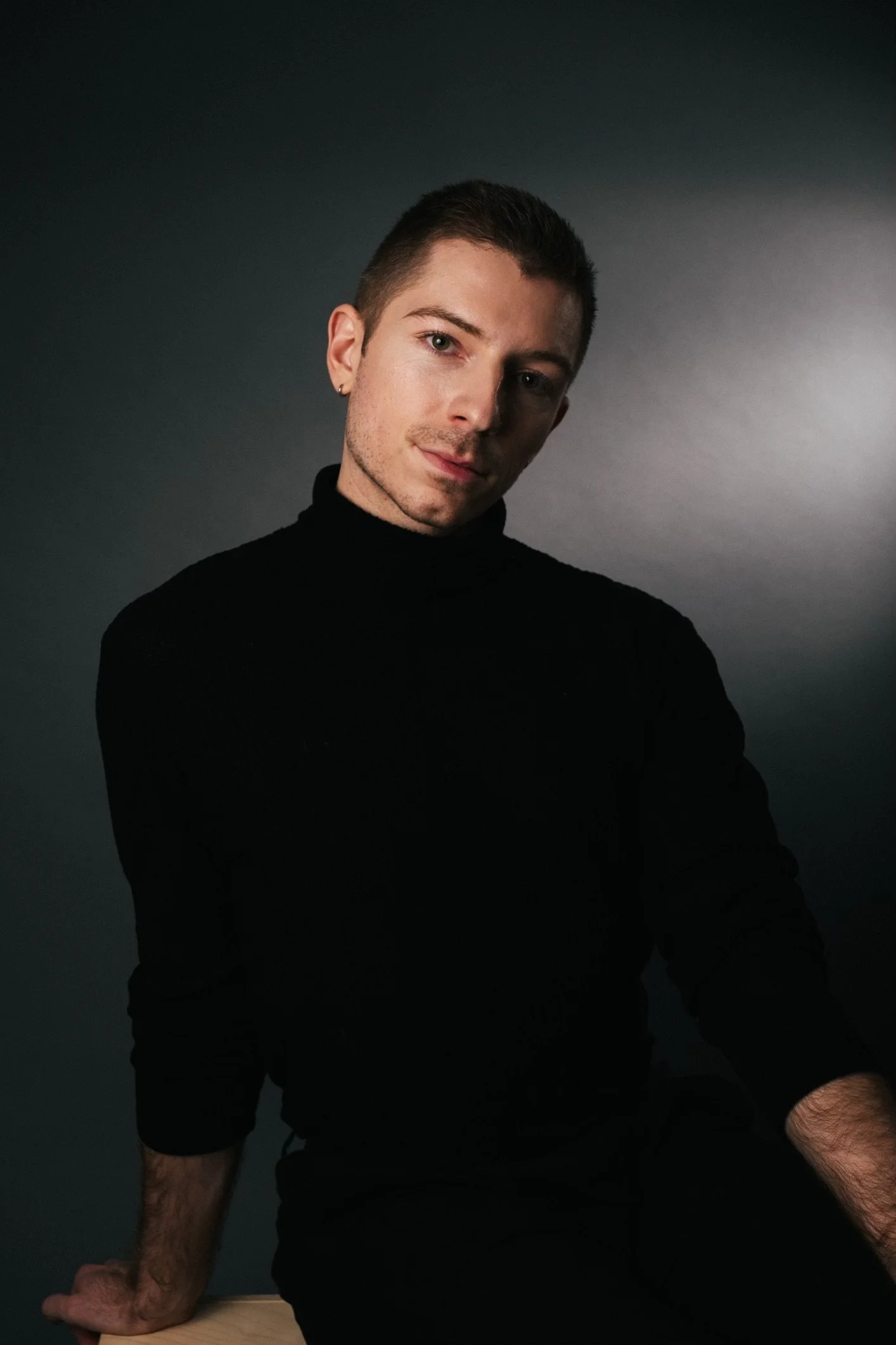 Portrait of a young man with short dark hair, light skin, wearing a black turtleneck, sitting against a dark gradient background.