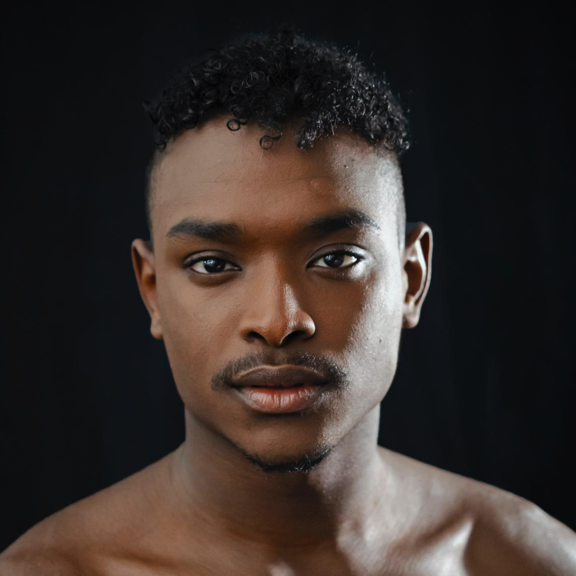 Close-up portrait of a young man with dark, curly hair, smooth skin, and a serious expression, against a black background.