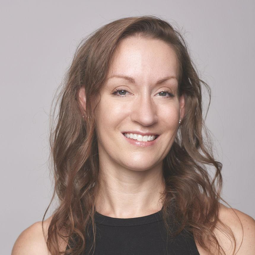 A woman with wavy brown hair smiling at the camera against a plain gray background.