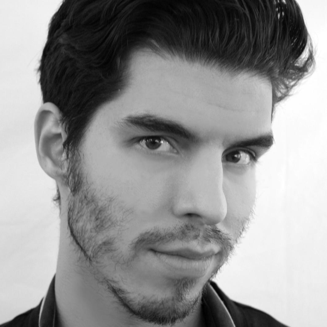 Black and white close-up portrait of a young man with wavy dark hair, a trimmed beard, and a slight smile.