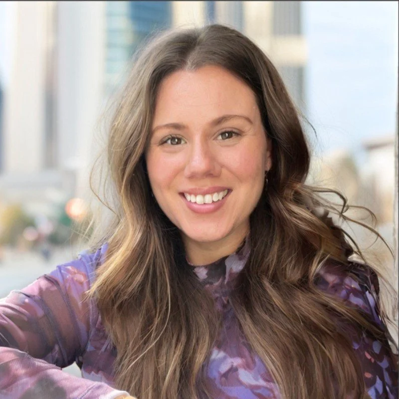 A woman with dark wavy hair wearing a white blouse is writing in a notebook with a black pen at a wooden desk.