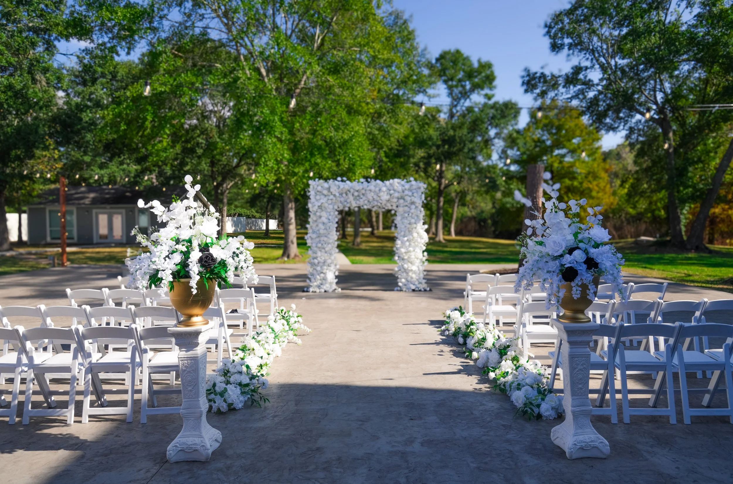 Outdoor wedding ceremony setup with white chairs, floral arrangements in gold vases, and a white flower arch under green trees on a sunny day.