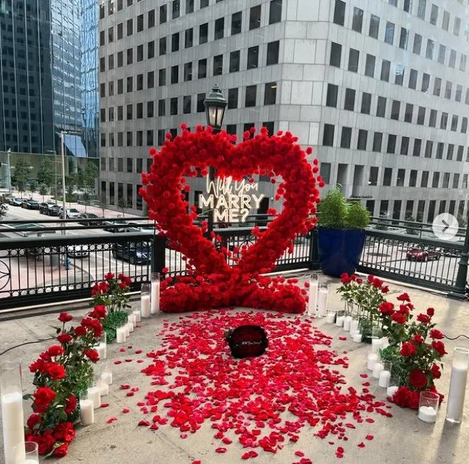 A romantic proposal setup on a balcony with a heart-shaped arrangement of red roses, white candles, and rose petals leading to a message board reading 'Will You MARRY ME?'