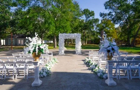 Outdoor wedding setup with white chairs, floral arrangements, and a floral arch in a park with trees