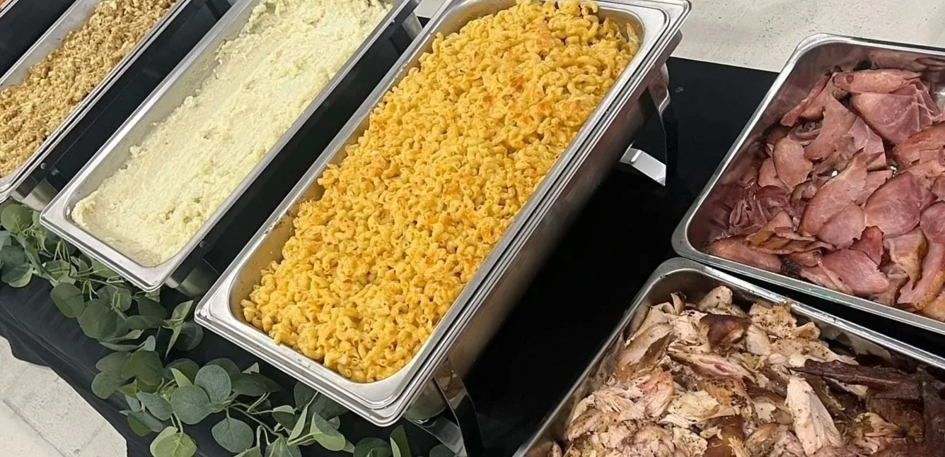 Buffet table with trays of pasta salads, sliced ham, and shredded chicken.