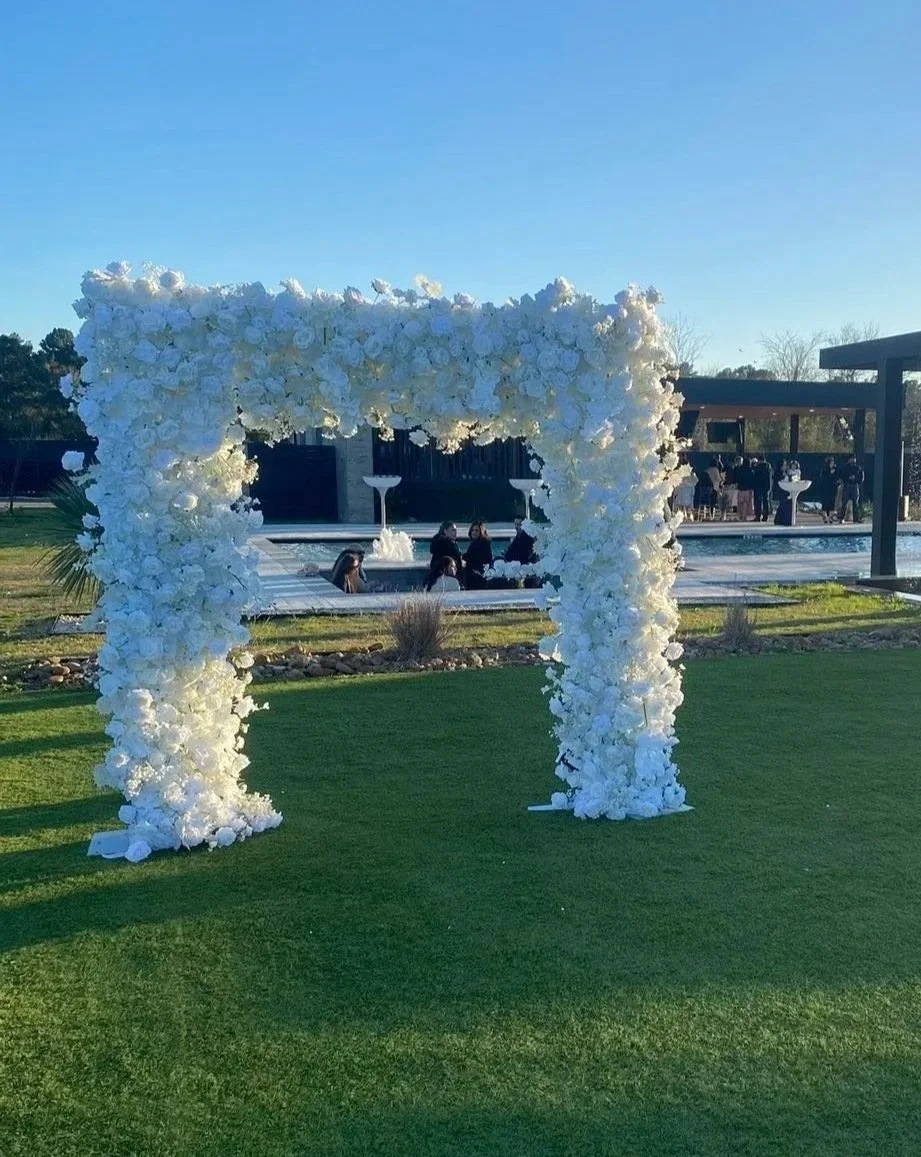 A large white flower arch on a lawn near a pool, with people in the background at an outdoor event during daytime.