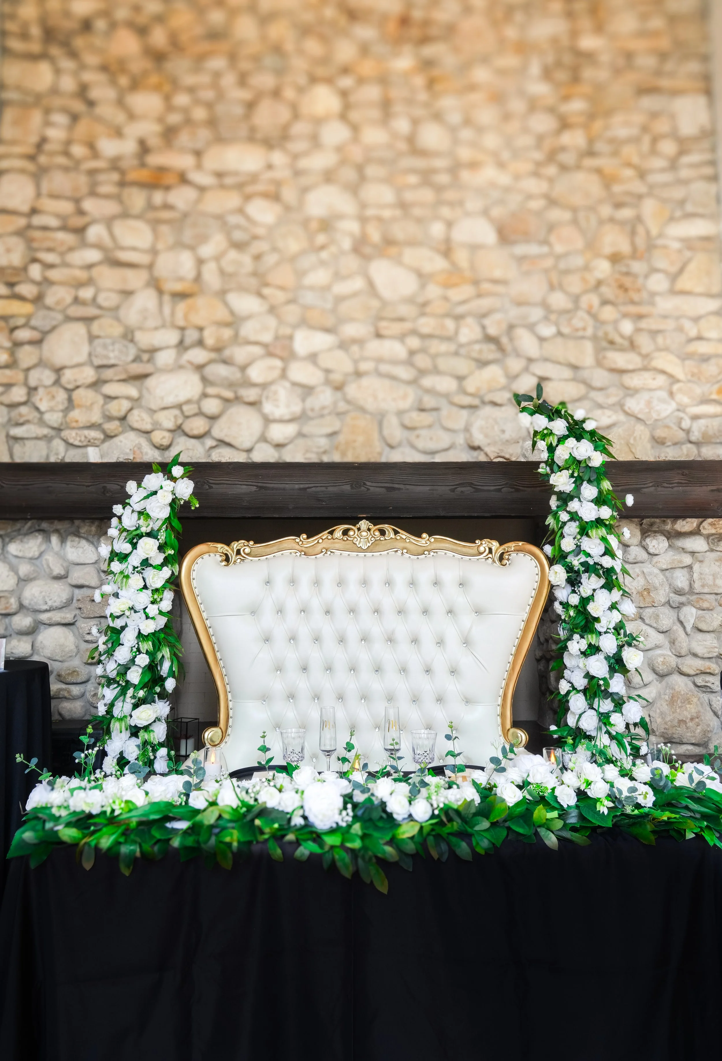 Elegant table with a white tufted headboard, decorated with white flowers and green leaves, set against a stone wall.