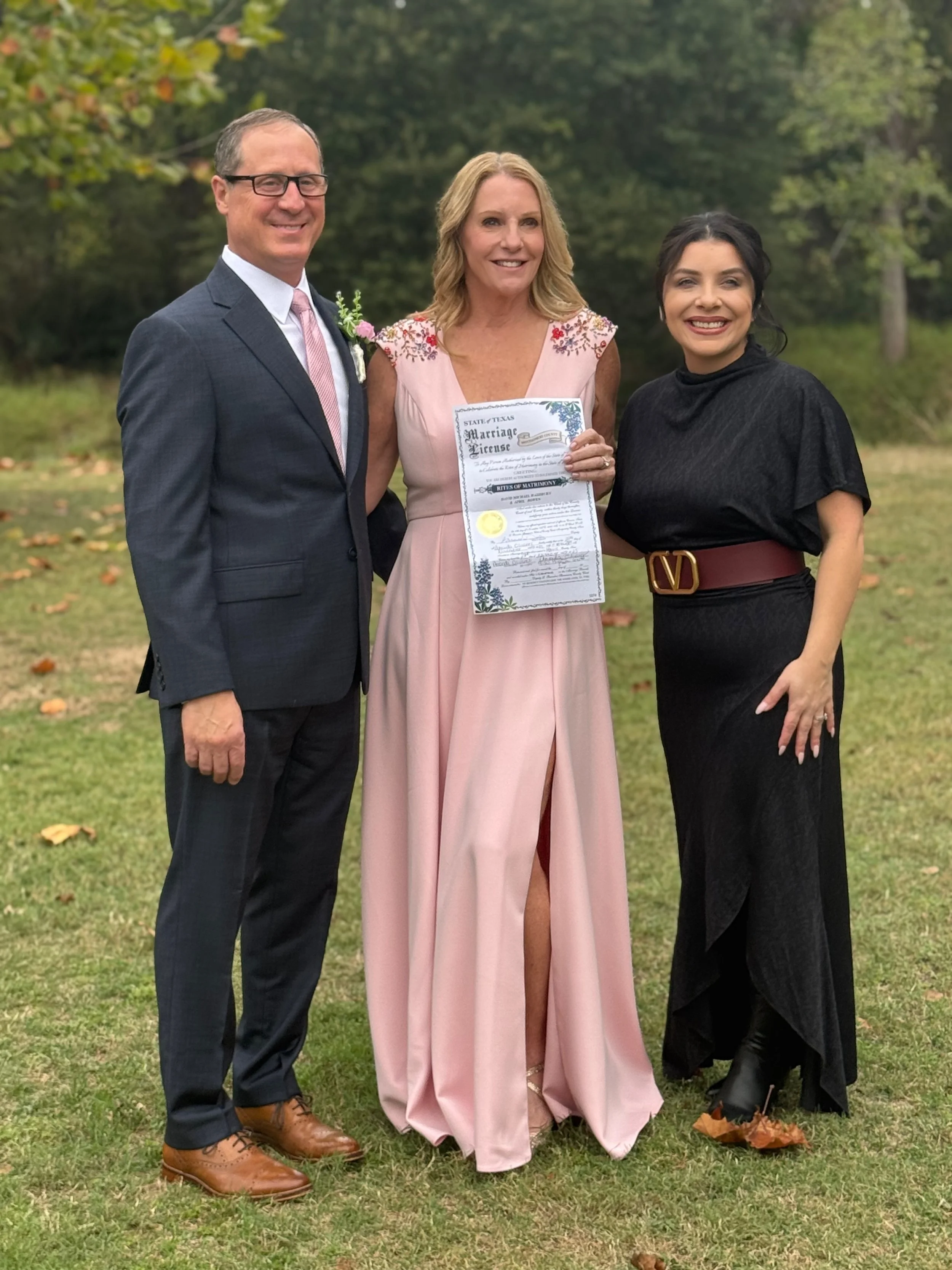 A woman in a pink dress holding a marriage license, flanked by a man in a suit and glasses on her left and a woman in a black dress with a red belt and black boots on her right, outside with green trees in the background.