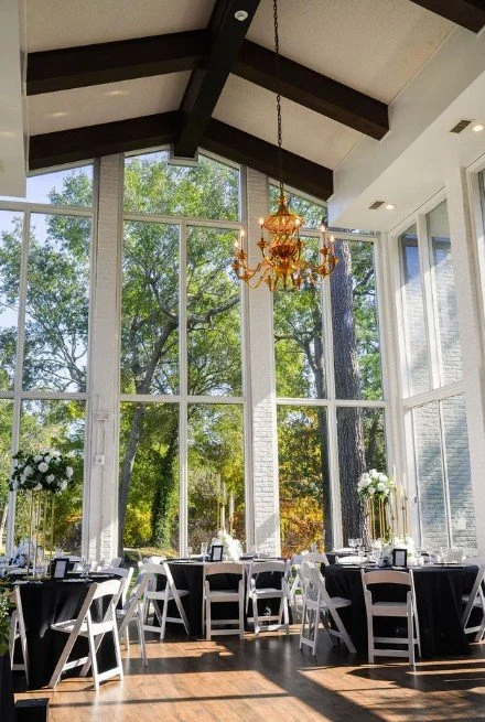 Elegant dining area with large floor-to-ceiling windows, black and white tables, white chairs, floral centerpieces, a gold chandelier, and exposed ceiling beams, with trees visible outside in natural light.