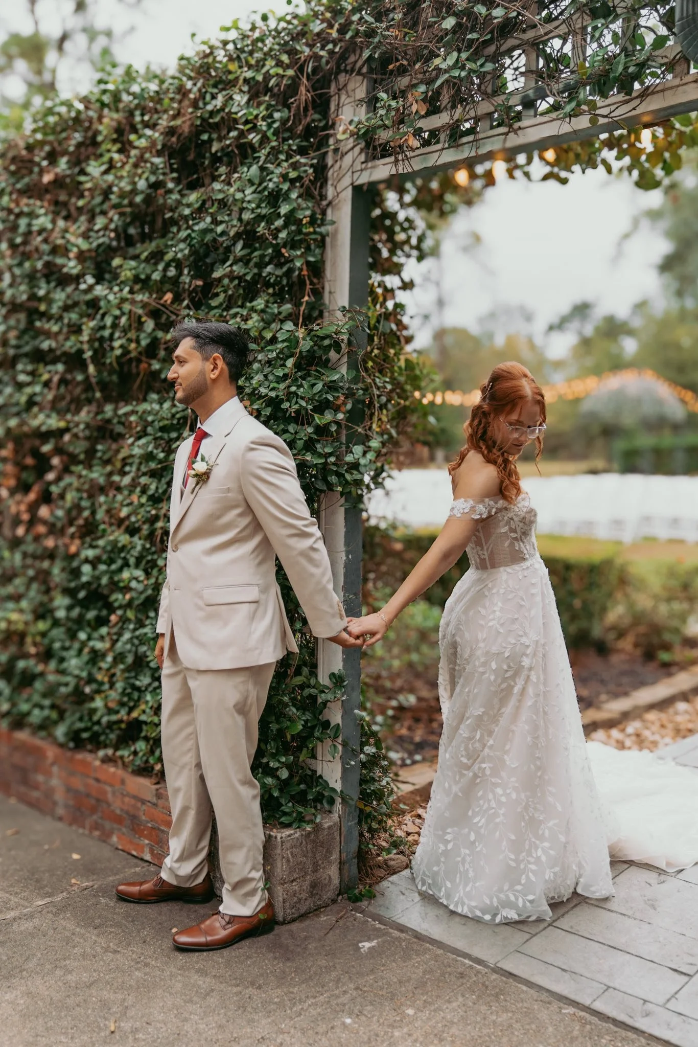 A bride and groom hold hands behind their backs, standing outdoors near a vine-covered arch at sunset during their wedding.