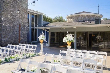 Outdoor event space with white chairs arranged in rows, floral arrangements in large vases, and a modern building with large windows in the background.