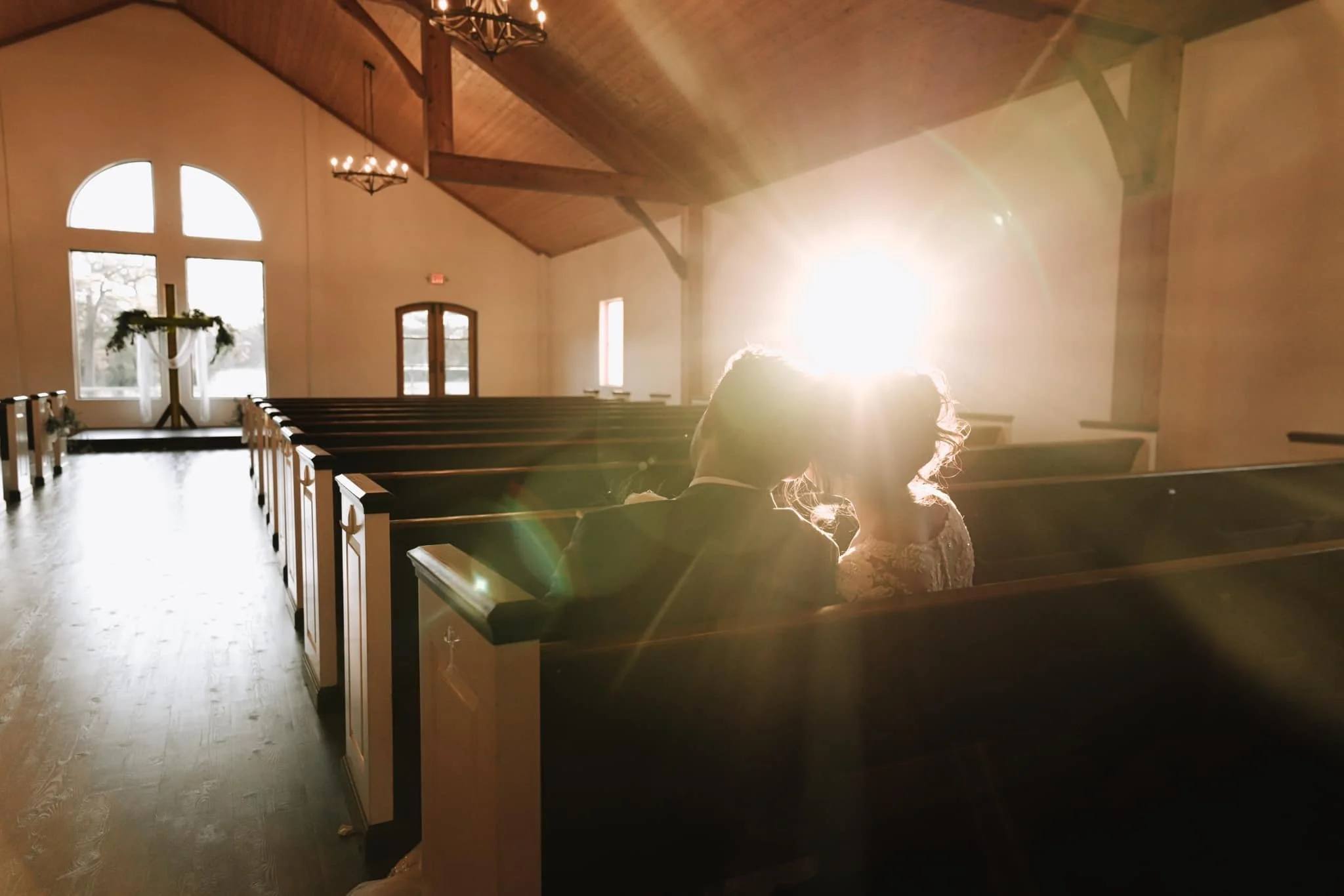 A romantic moment between a bride and groom sitting closely in a church with sunlight shining brightly behind them.