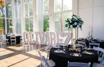 A decorated indoor event space with large windows, round tables set with plates and cutlery, and large illuminated letters spelling "OLIVAREZ" in the background.