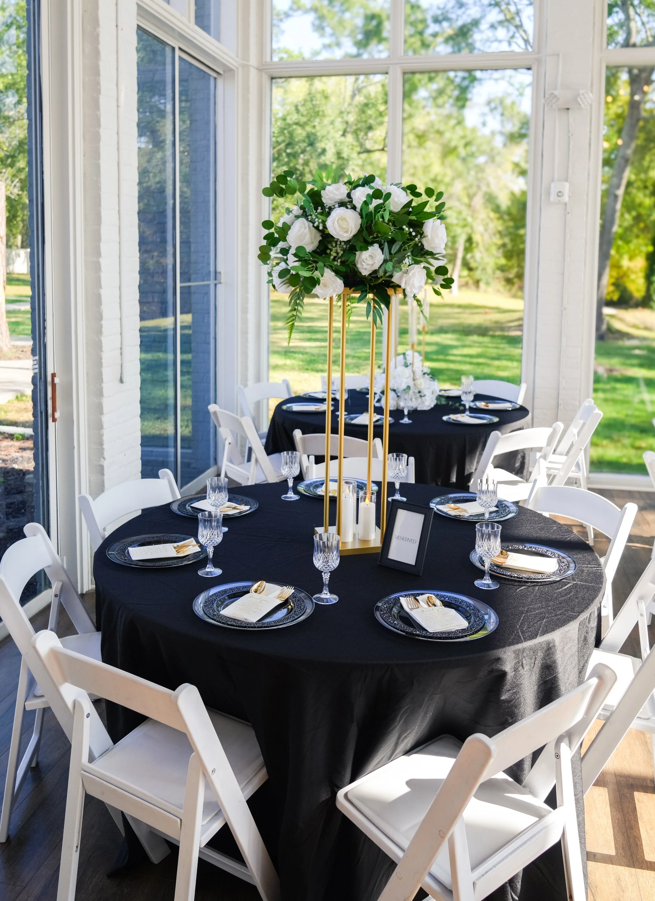 Round table decorated for a wedding reception with a tall floral centerpiece featuring white roses and greenery, set with black tablecloths, black and gold plates, crystal glasses, gold utensils, and reserved signs, inside a sunlit room with large wi