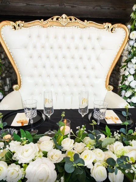 Elegant dining table set with white floral centerpiece, black placemats, champagne flutes, and gold accents, against a backdrop of a large ornate white and gold throne-like chair and white flowers.