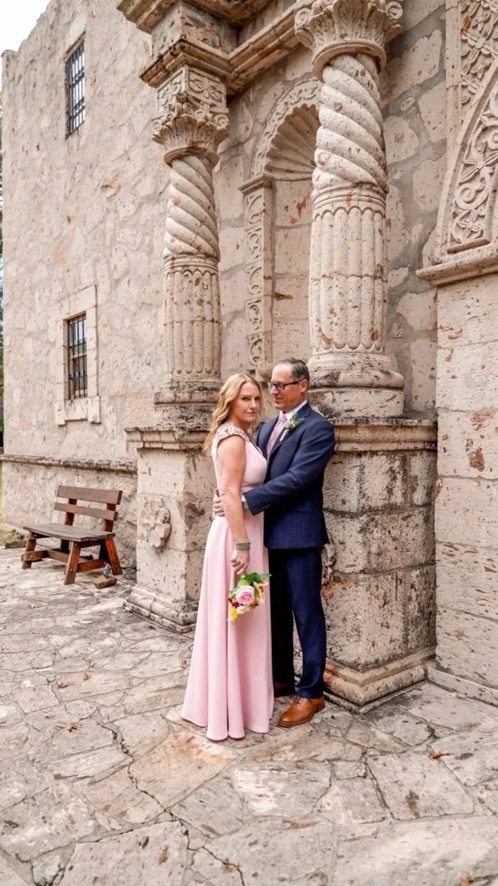A couple standing close together outside a historic stone building with ornate columns and arches, the woman holding a bouquet of pink and white flowers, both dressed formally for a special occasion.