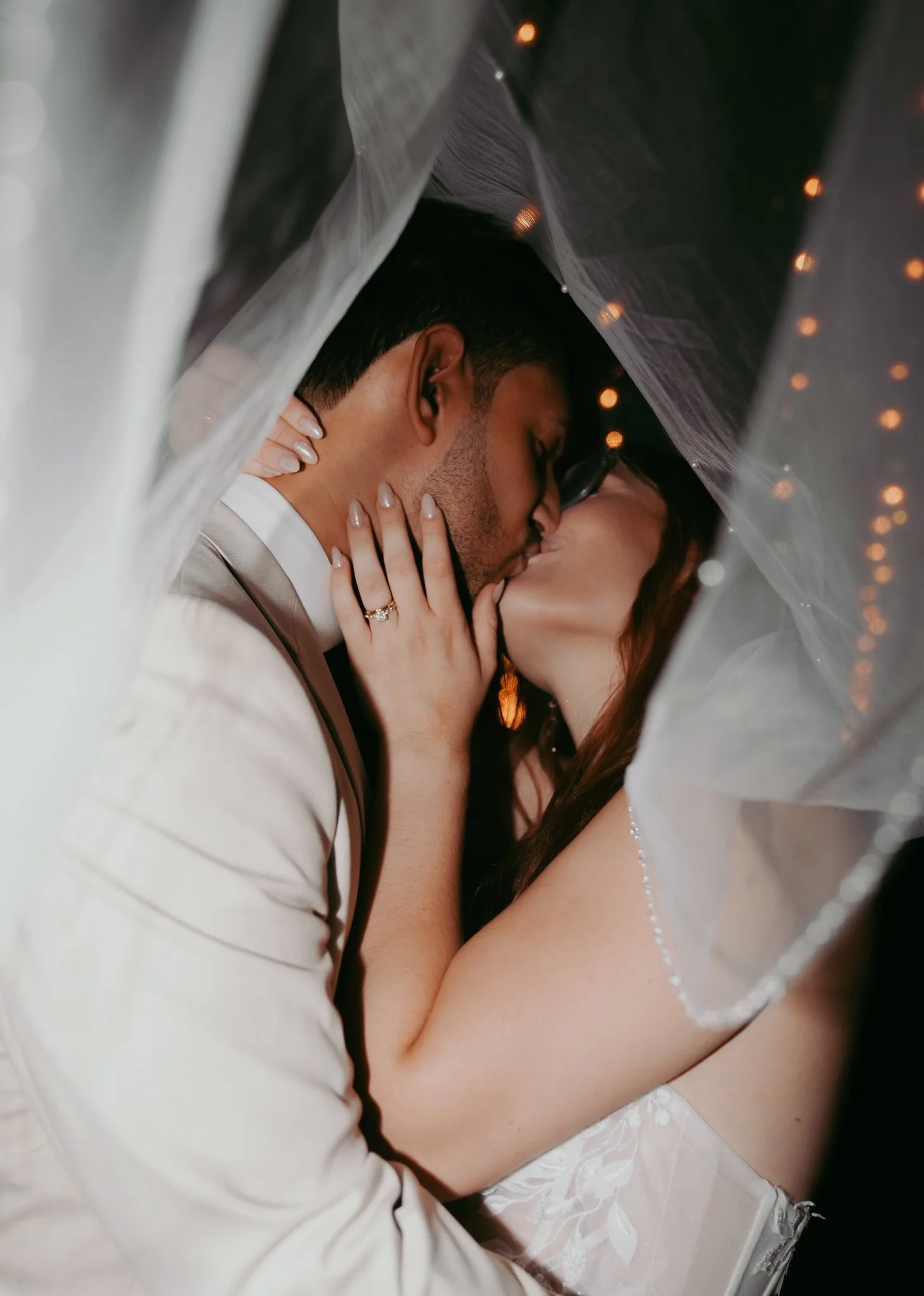 A bride and groom sharing a kiss under a veil, close-up shot showing their faces and hands, with wedding rings visible, during a wedding celebration.