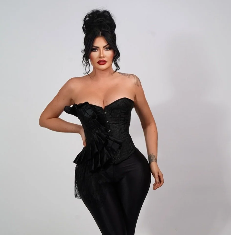 A woman with dark hair styled in an updo, wearing a black strapless corset, black pants, and bold makeup, standing against a plain light gray background.