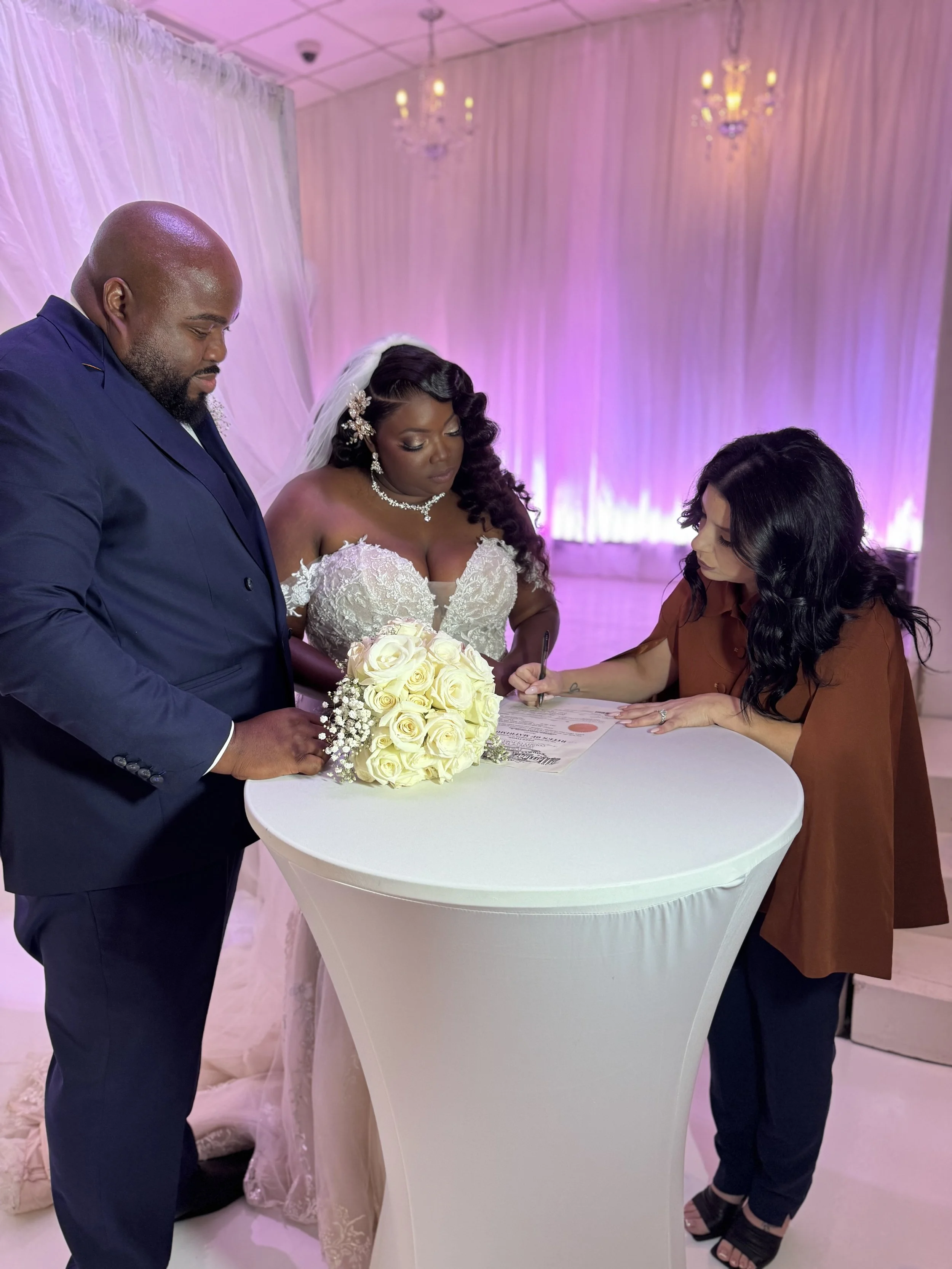 A bride and groom sign their marriage license with a woman assisting at a wedding reception, with a bouquet of white roses on a high white table, pink lighting, and elegant decor.
