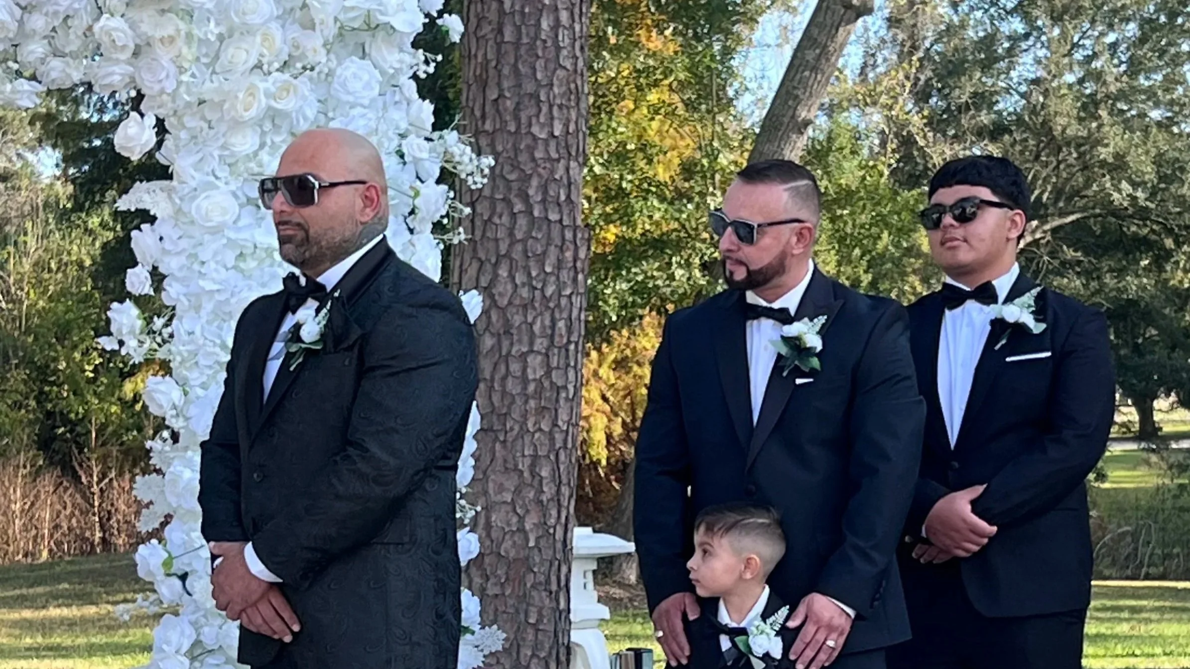 Three men in black tuxedos and sunglasses standing outdoors at a wedding ceremony, with a young boy in a tuxedo standing in front of them, under a floral arch of white roses and greenery, in a park-like setting with trees and grass.